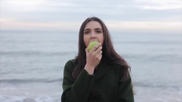 Sebuah close-up dari seorang gadis cantik mengenakan mantel hijau makan apel. Laut di latar belakang — Stok Video