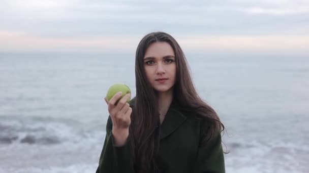 Un retrato de una hermosa niña feliz comiendo una manzana verde en un día fresco. Mar y amanecer cielo en el fondo — Vídeos de Stock