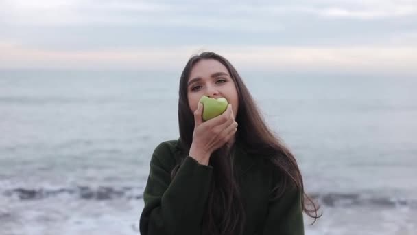 Um close-up de uma menina bonita em um casaco quente mastigar uma maçã verde na praia ao nascer do sol — Vídeo de Stock