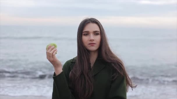 Movimento lento. Uma menina bonita em um casaco verde escuro comendo uma maçã à beira-mar ao nascer do sol. Close-up — Vídeo de Stock