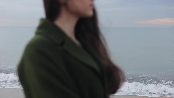 Een nauwe kant bijgesneden wazig zicht op een mooi meisje in een donker groene jas aan zee bij zonsopgang. De focus wordt verlegd van de achtergrond naar het meisje. Het meisje dat haar hand op haar schouder — Stockvideo