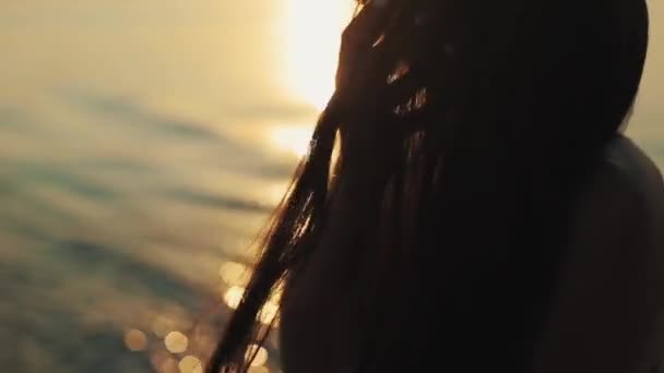 Una hermosa chica pasa sus dedos por su cabello mientras el sol se pone sobre el agua. Un primer plano. Una vista lateral — Vídeos de Stock