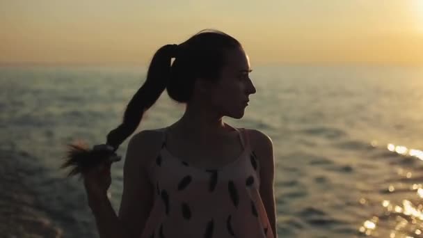 Un ritratto ravvicinato di una bella ragazza che guarda il lato e gioca con la coda di cavallo. Tramonto sul mare sullo sfondo — Video Stock