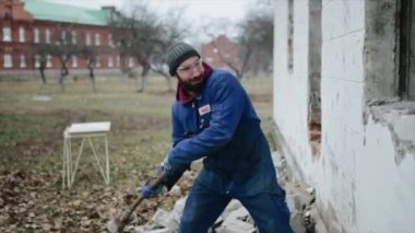 Bir işçi bir duvarı balyozla smashing bir yan görünüm. Binanın bazı bölümleri zaten yok edilir. Delikleri kare şeklinde. Yere tuğla.
