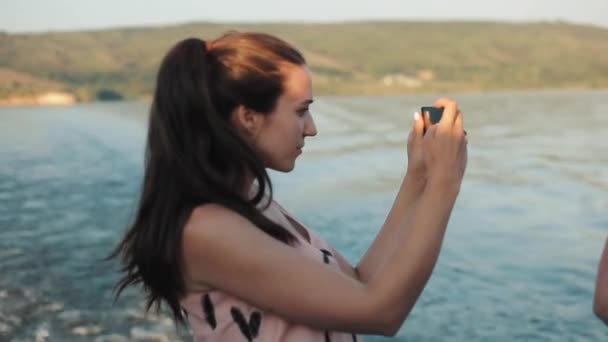 Вид сбоку на красивую девушку с хвостиком, фотографирующуюся на телефон с рекой и горами в солнечный день — стоковое видео