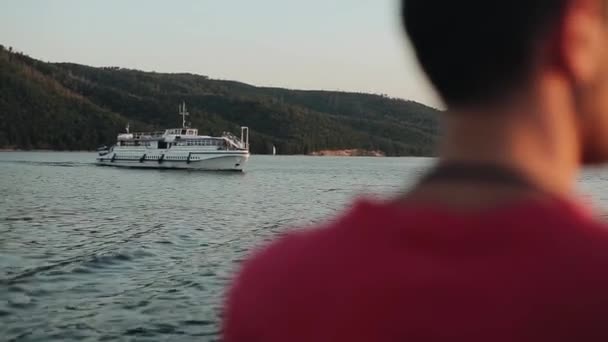 Вид ззаду на молодого чоловіка, який спостерігає за круїзним кораблем, що пливе вздовж річки. Гори і захід сонця небо на задньому плані — стокове відео