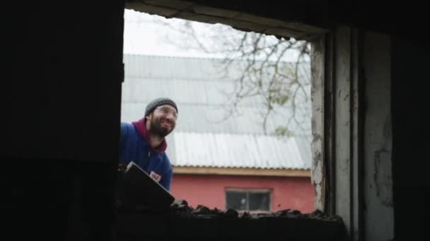 Mit dem Vorschlaghammer eine Mauer durchschlagen. Fallende Ziegel. Ein Arbeiter mit Schutzbrille und blauem Overall. Blick von innen. — Stockvideo