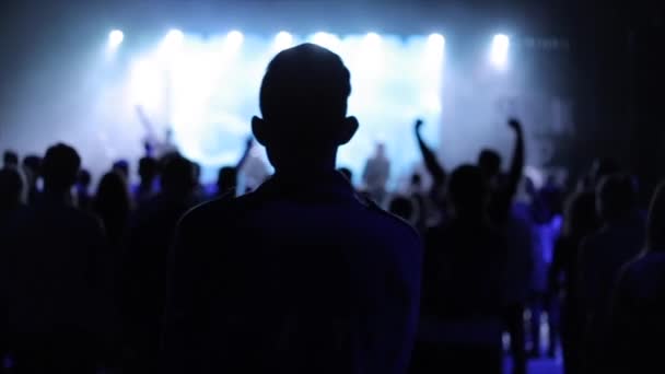 Vista trasera de un hombre en un concierto escuchando música lenta. Mucha gente está frente a él. Algunos de ellos están levantando las manos. Adoración — Vídeo de stock