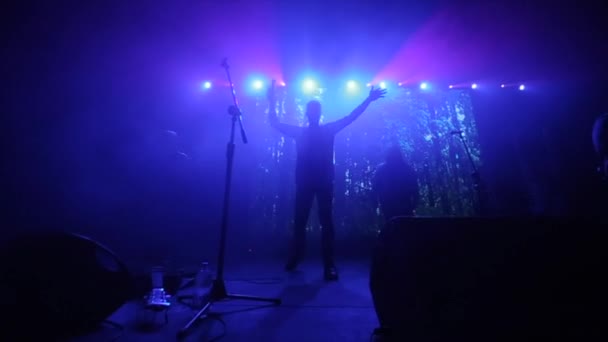 Ein Mann mit zwei erhobenen Händen bei einem Konzert. Blaulicht. Rockband. die Frau, die im Hintergrund springt — Stockvideo