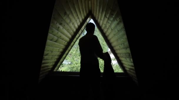 O vedere din spate a unui bărbat care pune o jachetă în fața unei ferestre triunghiulare într-o cameră de mansardă din lemn — Videoclip de stoc