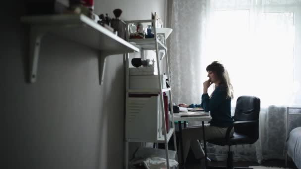 Vue latérale d'une belle jeune fille assise à la table de travail, tenant son téléphone portable — Video