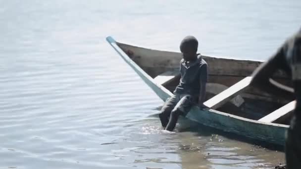 Kenia, Kisumu - 20 mei 2017: Mooie Afrikaanse jongen is zitten in de boot op de oever van het meer en zijn benen slingeren. — Stockvideo