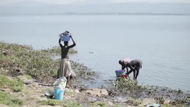 Кенія, Кісуму - 20 травня 2017: Жінка і її дочка мити посуд у озеро, дівчина нести басейну з одягом на голову. — стокове відео