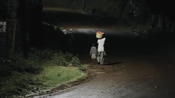 Baksidan på afrikanska mannen bär korg med kläder, två pojkar som går nära hane genom vägen på natten i regn. — Stockvideo