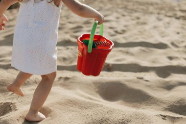 Kırmızı bir kova ile beyaz elbiseli plaj boyunca kaçan küçük beyaz kız. Arkadan görünüm