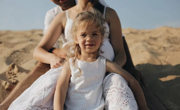 The little beautiful caucasian girl standing between her parents legs. Pregnant mother. Happy family in whote clothes stand on the sand Royalty Free Stock Images