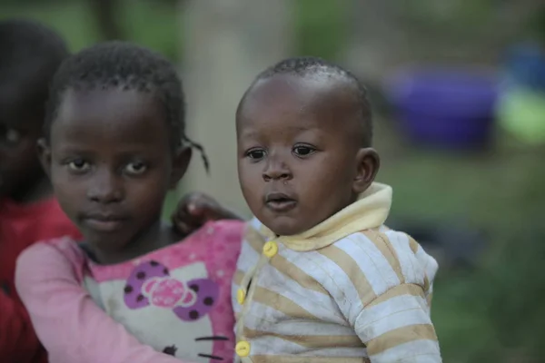 KENIA, KISUMU - 20 DE MAYO DE 2017: Vista trasera de una niña africana llevando a un bebé desnudo por el pueblo . — Foto de Stock