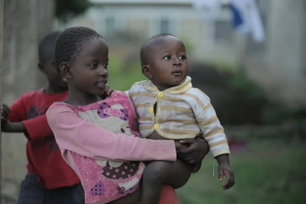 KENYA, KISUMU - MAY 20, 2017: Back view of little african girl carrying naked baby boy through the village. Stock Image