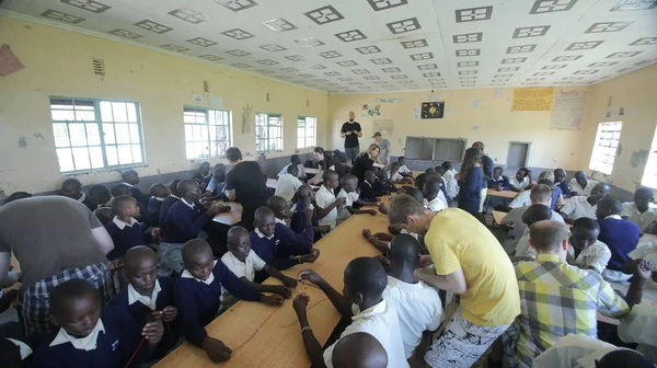 KENYA, KISUMU - MAIO 23, 2017: Grande grupo de crianças e adolescentes africanos sentados, voluntários caucasianos ajudam a fazer pulseiras — Fotografia de Stock