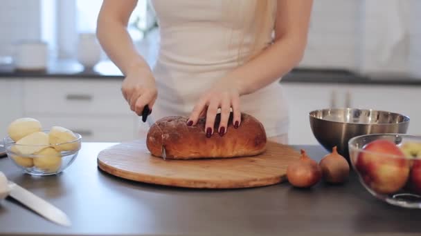 Closeup γυναικεία χέρια κοπή ψωμιού. Το μαγείρεμα στην κουζίνα. Καυκάσιος κορίτσι σε λευκό πουκάμισο με τα κόκκινα νύχια. Ελαφριά κουζίνα. Όμορφο φρέσκο ψωμί. — Αρχείο Βίντεο
