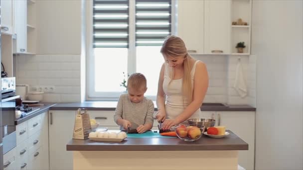 Una joven hermosa madre caucásica con el pelo blanco y el hijo cocinan en una cocina brillante. Mamá enseña a hijo a cocinar . — Vídeos de Stock