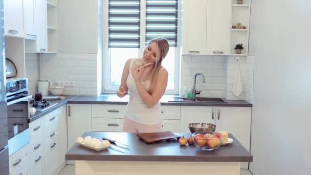 Beautiful caucasian girl in white shirt and pink cooking in the light kitchen, talking on the mobile phone and smiling while cooking in kitchen at home. — Stock Video