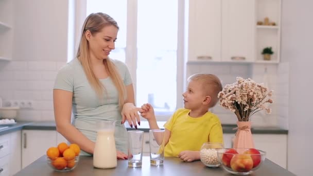 Beautiful caucasian mom fooling around with her son in the kitchen at the table. Family having fun at the kitchen. Colorful bright shirt. Beatiful light modern kitchen. Bright shirts. — Stock Video