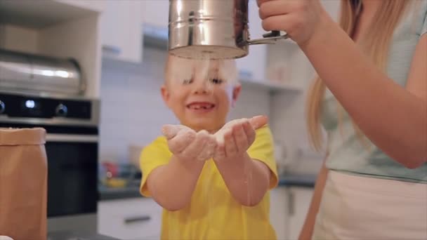 Belle maman caucasienne verse de la farine sur les mains de son fils. Un petit garçon aux cheveux blancs et aux grands yeux en chemise jaune rit et s'amuse . — Video