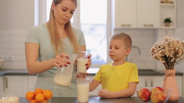 Piękna Mama kaukaski z długie, białe włosy, wylewanie szklankę mleka dla syna Ładna w żółte koszulki. Nowoczesna kuchnia. Siedzi na stole. Rodzinne śniadanie. Owoce i kwiaty na stole — Wideo stockowe