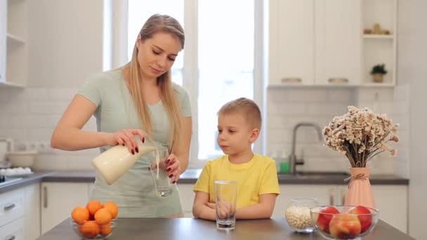 Όμορφη μαμά Καυκάσιος με μακριά λευκά μαλλιά ξεχειλίζει ένα ποτήρι γάλα για το χαριτωμένο γιο σε κίτρινο πουκάμισο. Μοντέρνα κουζίνα. Κάθεται στο τραπέζι. Οικογενειακών προγευμάτων. Φρούτα και λουλούδια στο τραπέζι — Αρχείο Βίντεο