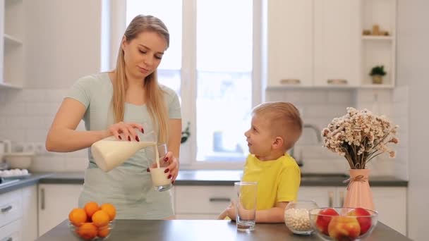 美丽的高加索妈妈与长的白发倾吐出一杯牛奶为她可爱的儿子在黄色衬衫。现代化的厨房。坐在桌子上。家庭早餐。桌子上的水果和鲜花。儿子 — 图库视频影像