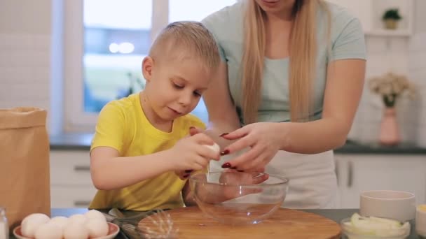 Mooie caucsian kind met blauwe ogen bakken voorbereiden taart. Moeder jonge vrouw onderwijs tonen van haar zoon kind hoe breken ei in kom. Moderne keuken. — Stockvideo