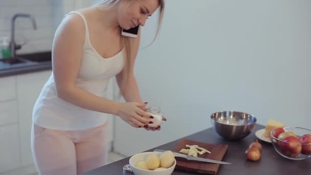 Beautiful girl is mixing food in frying pan, talking on the mobile phone and smiling while cooking in kitchen at home — Stock Video