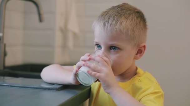 Closeup Καυκάσιος αγόρι με μεγάλα μπλε μάτια που πίνει γάλα στην κουζίνα. Παιδί σε κίτρινο πουκάμισο. Αγόρι χαμογελά στη φωτογραφική μηχανή. η μητέρα σκουπίζει το στόμα τους γιους. — Αρχείο Βίντεο