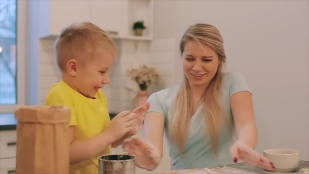 Schattige kleine jongen en zijn mooie moeder in kleurrijke shirtjes spelen en lachen tijdens het kneden van het deeg in de keuken. Moeder en zoon spelen met bloem — Stockvideo