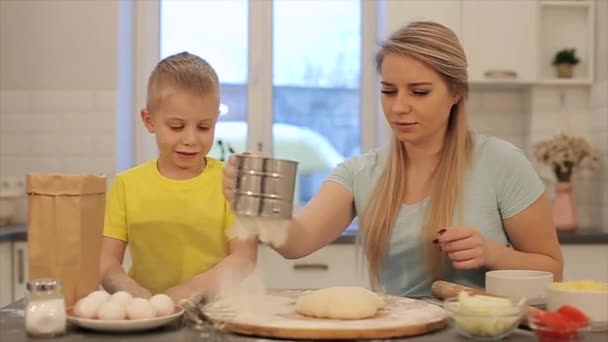 Vista frontal do menino bonito e sua bela mãe em camisas coloridas estão brincando e rindo enquanto amassa a massa na cozinha. Mãe e filho brincam com farinha — Vídeo de Stock