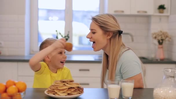 Маленький белый блондин веселится с молодой мамой. Мальчик дразнит маму блинчиками. Счастливая семья завтракает вместе на кухне . — стоковое видео