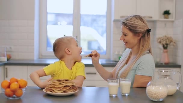 利特高加索金发碧眼的男孩与年轻的妈妈有乐趣。年轻的母亲戏弄男孩煎饼。愉快的家庭一起在厨房里吃早餐. — 图库视频影像