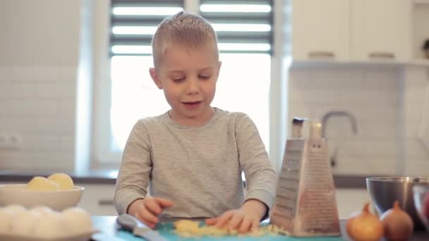 Little beautiful caucasian boy with big blue eyes cooking in the bright kitchen. He is pours cheese. Cheese grater. Happy cheerful kid make pizza. — Stock Video