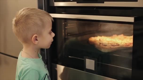 Little boy in blue shirt watching as pizza prepares in oven indoors. — Stock Video