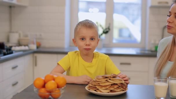小白种人与大蓝色眼睛在黄色衬衫有乐趣在厨房里。早上。儿子和母亲在一起现代厨房。坐着的男孩。带煎饼的男孩早上吃早餐。厨房里的灰色桌子。年轻 — 图库视频影像
