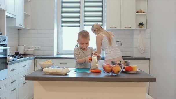 Little handsome boy rubs a carrot at the table. A young beautiful caucasian mother with white hair and son cook in a bright kitchen. — Stock Video