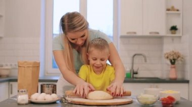 Küçük güzel sarışın çocuk annesi pişirmek için yardımcı olur. Anne ve oğlu renkli gömlek içinde dışarı hamur rulo vardır. Anne arkasında duruyor.