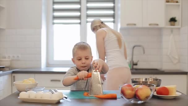 ハンサムな男の子はテーブルでニンジンをこすり。若い美しい白い髪と息子を持つ白人母明るいキッチンで調理します。. — ストック動画