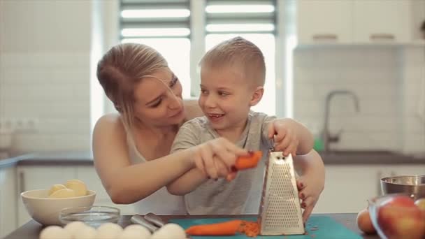 Mom teaches son to rub carrot. They are laughing. A young beautiful mother with in white shirt and cute son cook in a white kitchen.