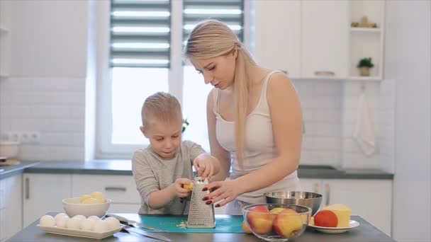 Μαμά διδάσκει ο γιος να τρίβετε το τυρί. Μια νεαρή όμορφη μητέρα με λευκό πουκάμισο και χαριτωμένο γιος μάγειρας σε μια άσπρη κουζίνα. — Αρχείο Βίντεο