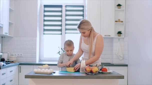 A mãe ensina o filho a cortar queijo. Uma jovem mãe bonita com camisa branca e bonito filho cozinhar em uma cozinha branca . — Vídeo de Stock