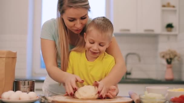 De kleine mooie blonde jongen helpt moeder om te koken. Moeder en zoon in kleurrijke shirtjes zijn het deeg uitrollen. Mam staat achter. — Stockvideo