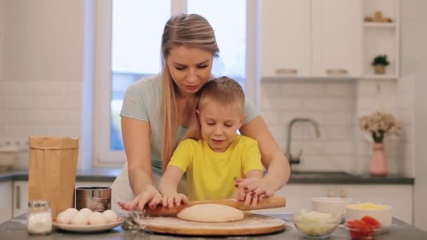 Beautifull blond pojken hjälper mamma att laga mat. Mamma och son i färgglada skjortor är Kavla ut degen. Mamma står bakom. — Stockvideo