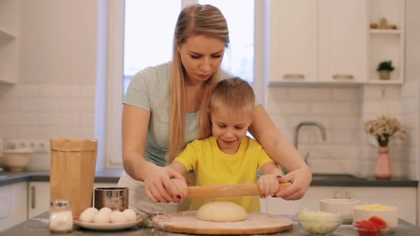 De kleine mooie blonde jongen helpt moeder om te koken. Moeder en zoon in kleurrijke shirtjes zijn het deeg uitrollen. Mam staat achter. Moderne keuken. — Stockvideo